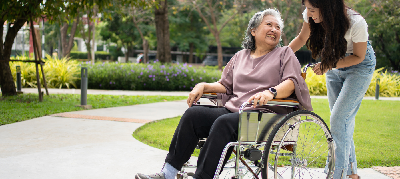 Une femme pousse une femme en fauteuil roulant dans un parc. Elles se sourient.